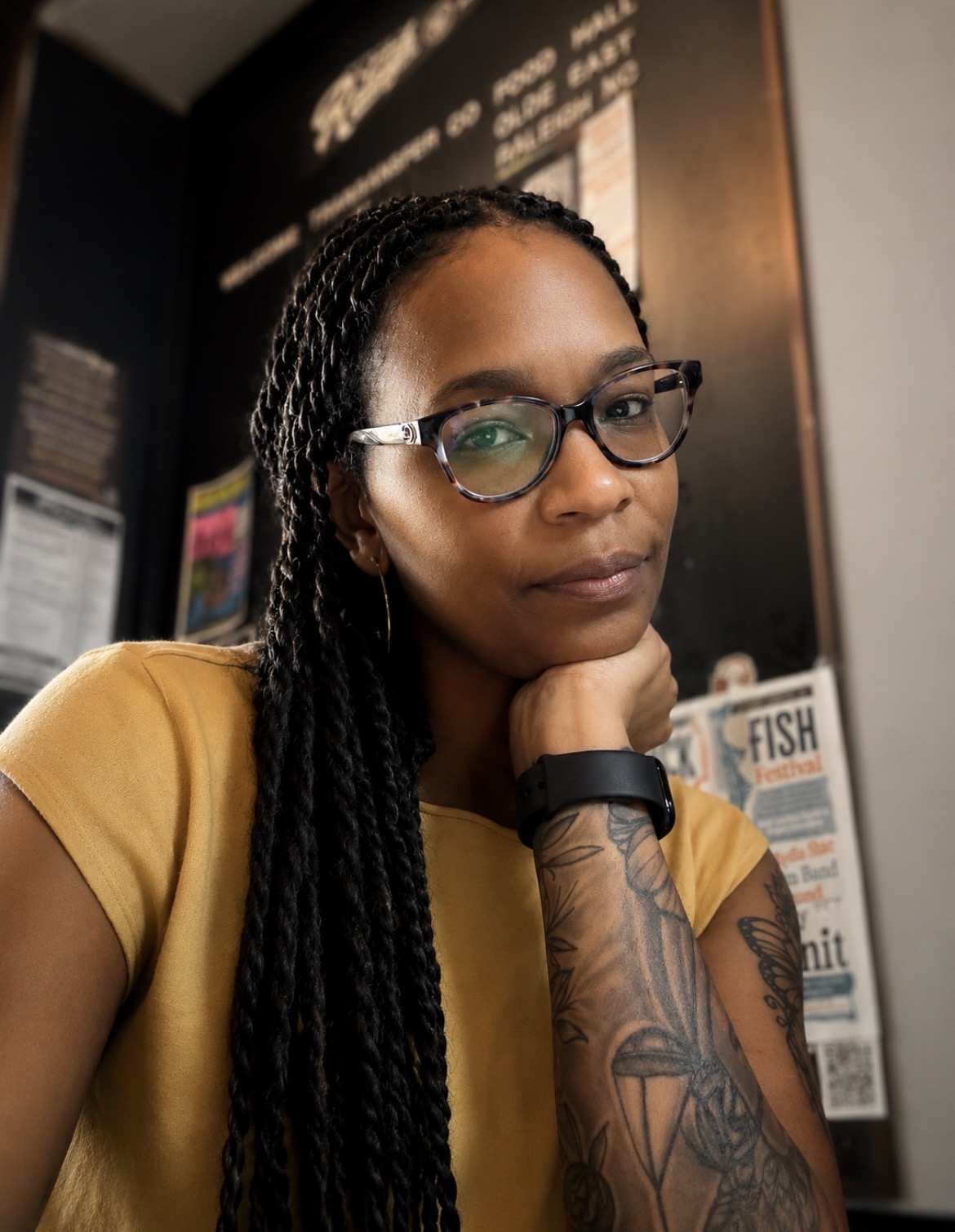 Woman with glasses and long braided hair resting her chin on her hand, wearing a yellow top, in an indoor setting.