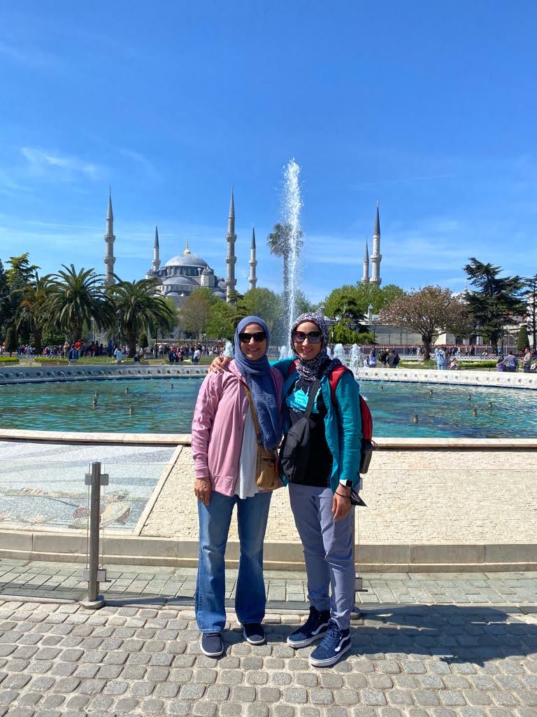Two women stand in front of a fountain with a historic mosque and minarets in the background, under a clear blue sky.