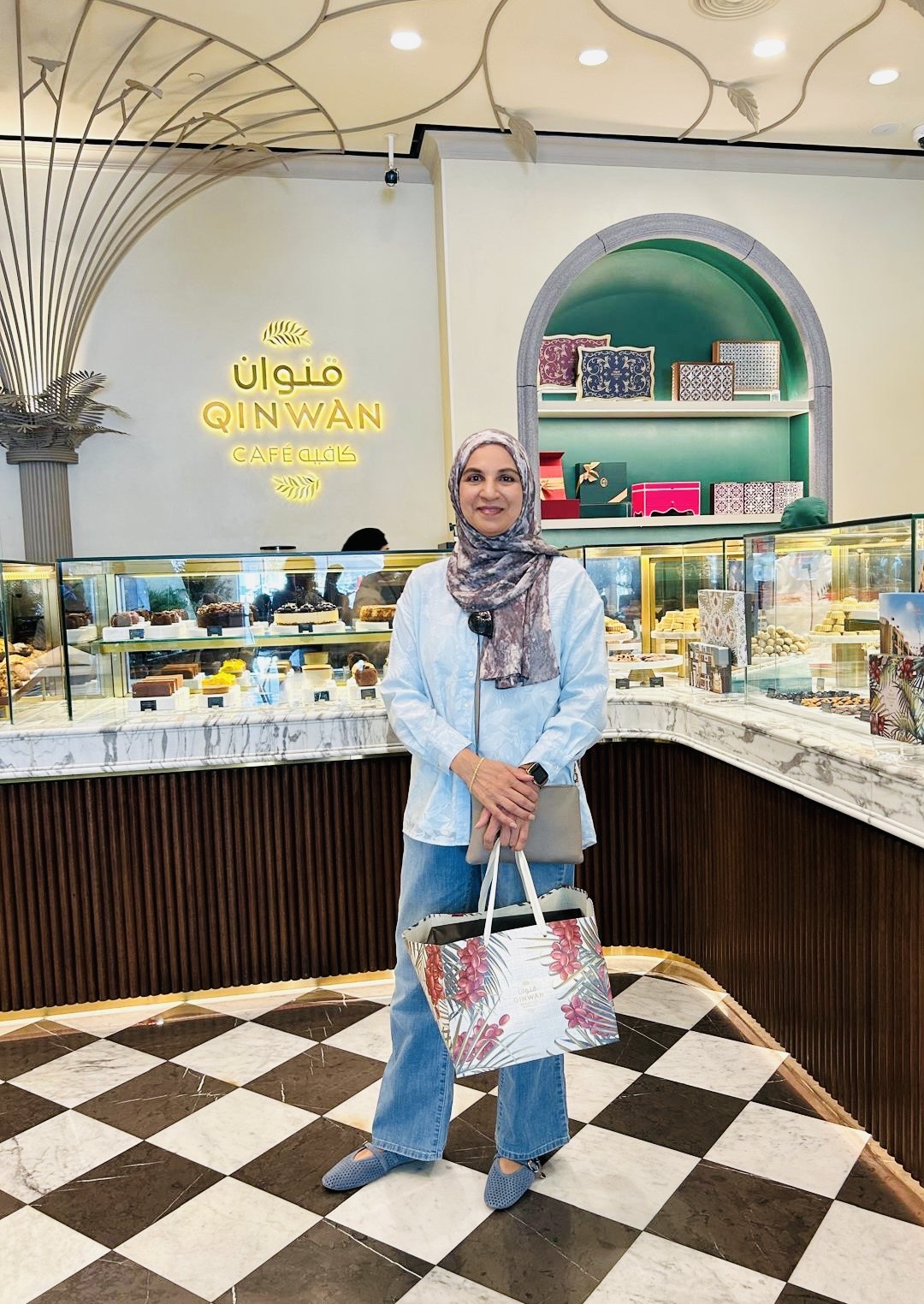 Woman wearing a hijab and blue outfit standing in a bakery with a display case and decorative interior.