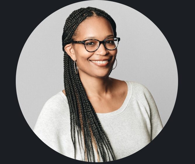 Smiling woman with glasses and braided hair, wearing a light-colored top, against a gray background.