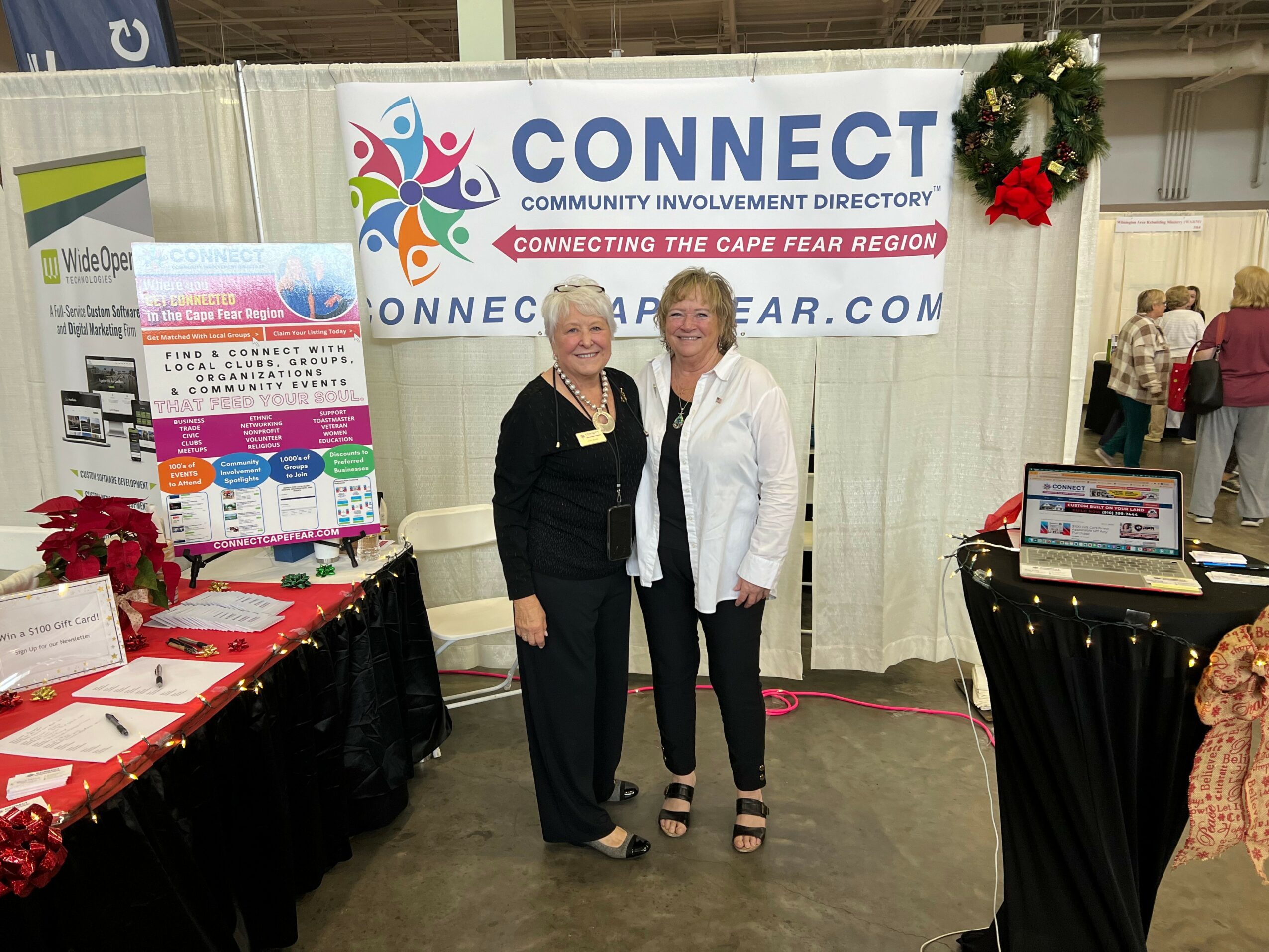 Two women standing in front of a banner that reads 'CONNECT' at an event booth, with tables displaying pamphlets and decorations.