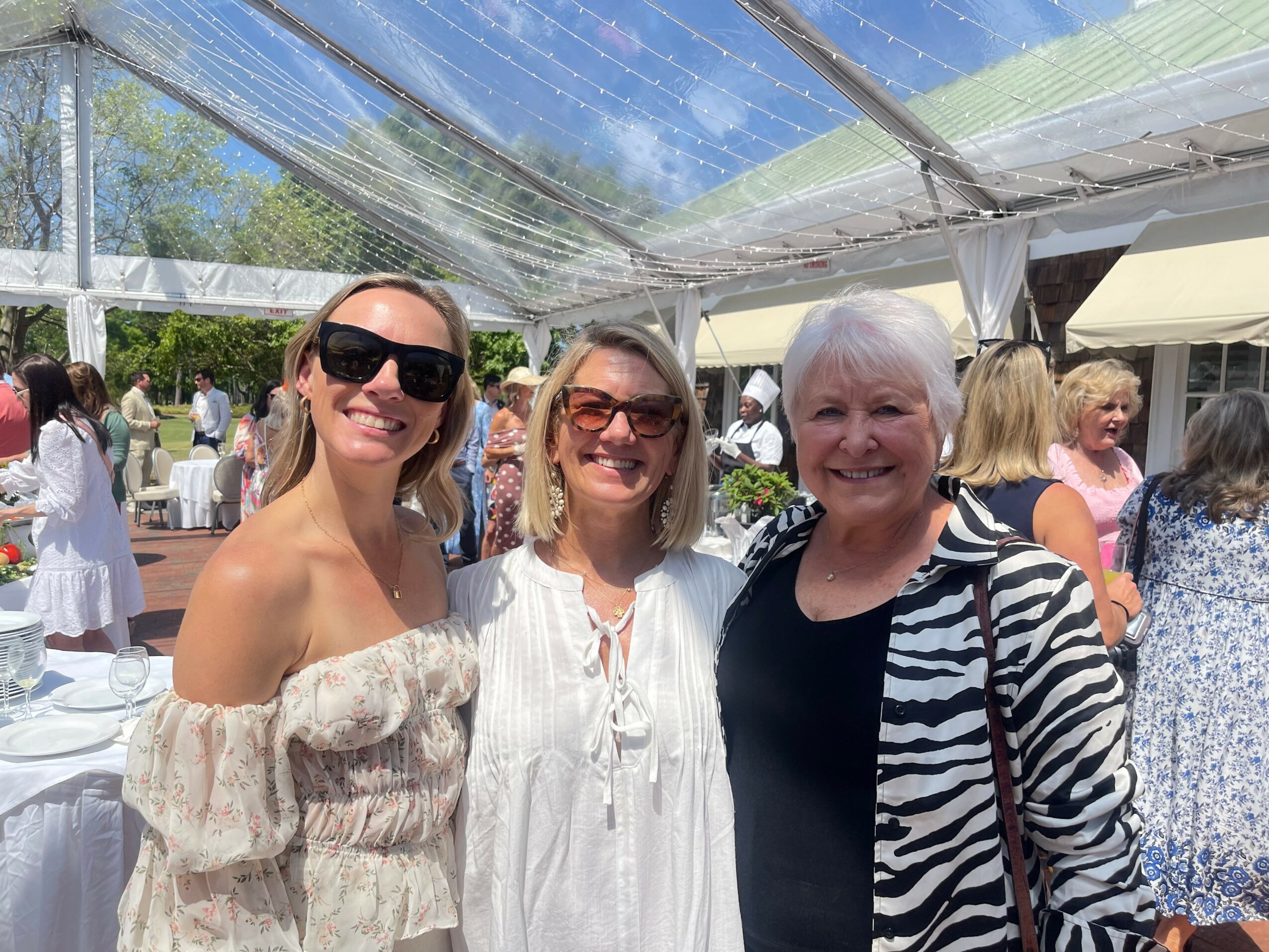 Three women smiling outdoors under a transparent tent, with other people in the background, sunny day.
