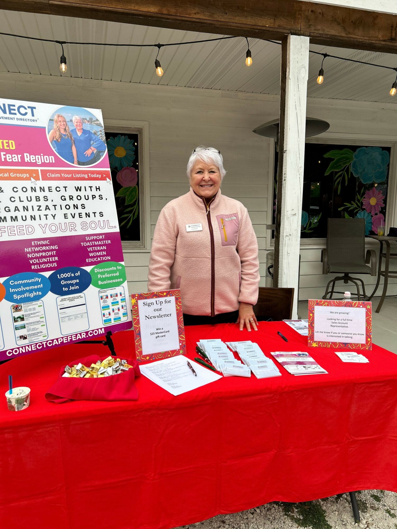 Woman standing behind a table with informational materials, in an indoor space with colorful artwork on the wall.