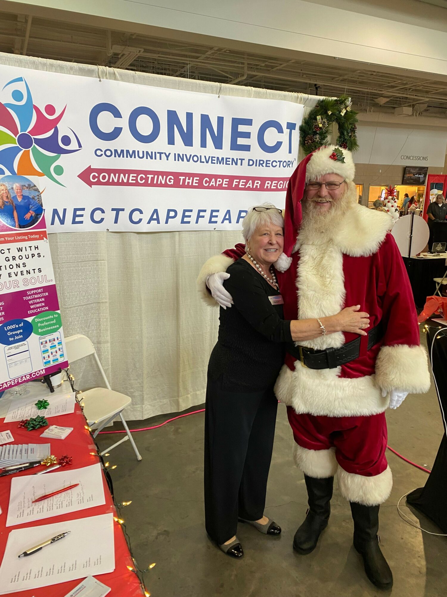Person dressed as Santa Claus standing with a woman in front of a banner, both smiling, in an indoor setting.