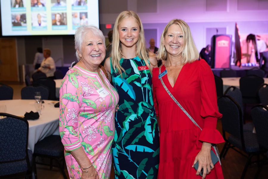 Three women smiling at an event, standing close together in a decorated room with tables and chairs.