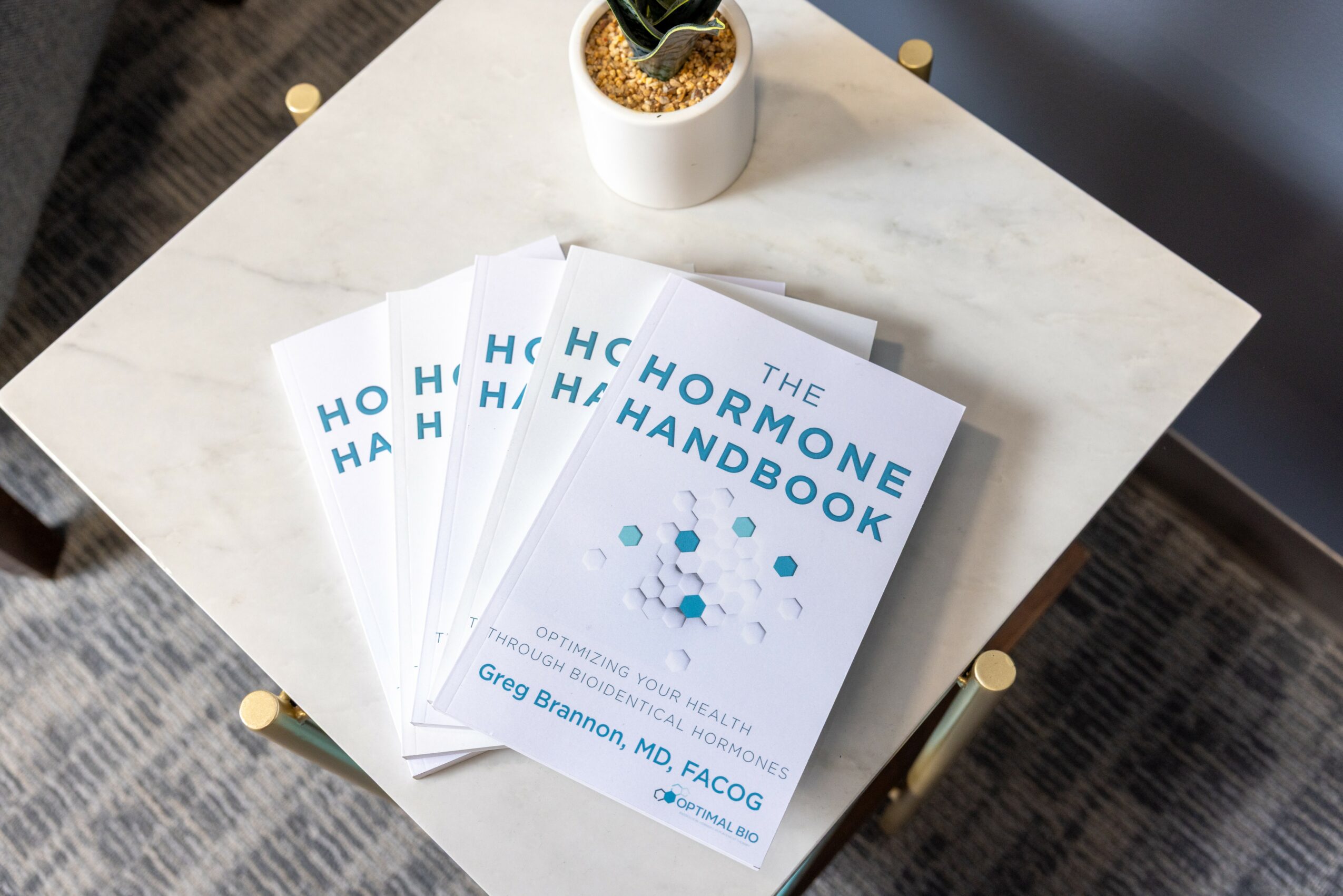 Five copies of 'The Hormone Handbook' book stacked on a white table with a potted plant nearby.