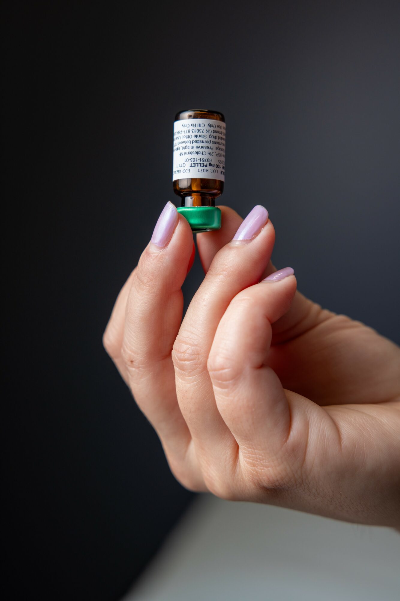 Hand holding a small brown vial with a white label and green cap against a dark background.