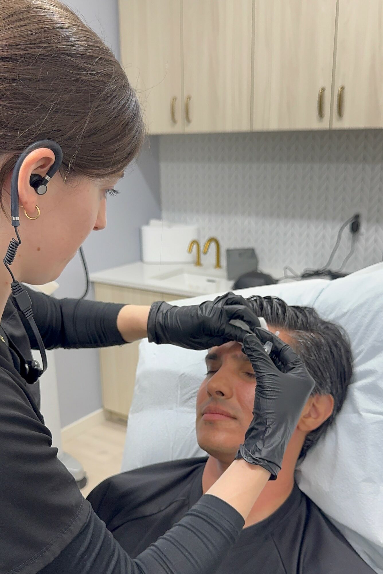 Medical professional in black gloves examines a woman's face with a tool, in a clinical setting with cabinets and sink.