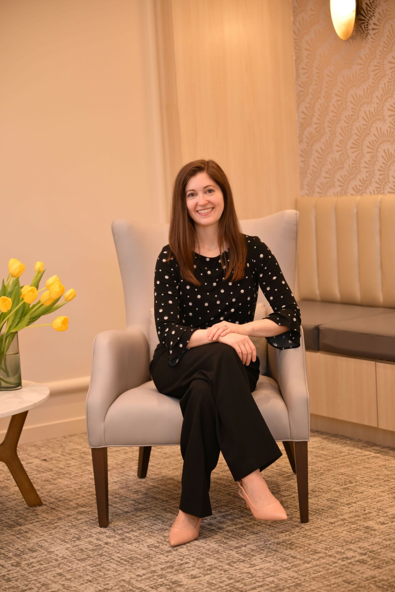 Woman sitting on a chair in a room with a table and flowers, smiling, wearing a black polka dot top and black pants.