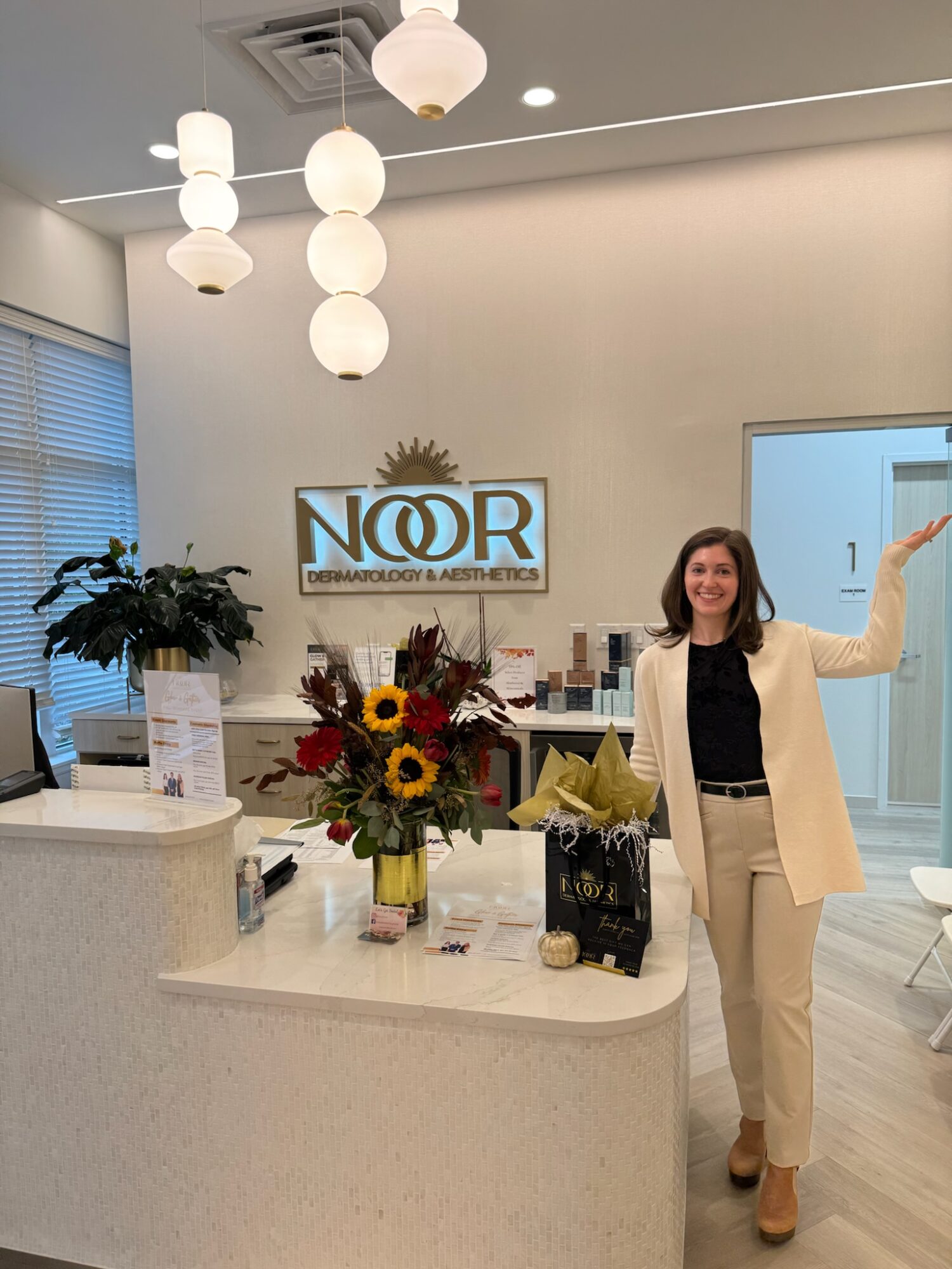 Woman standing behind reception desk, smiling, waving, with flowers and office sign in background.
