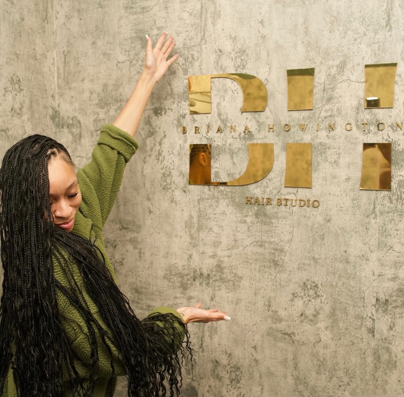 Woman with long curly hair raises one arm, standing next to a wall with gold letters. Neutral background.