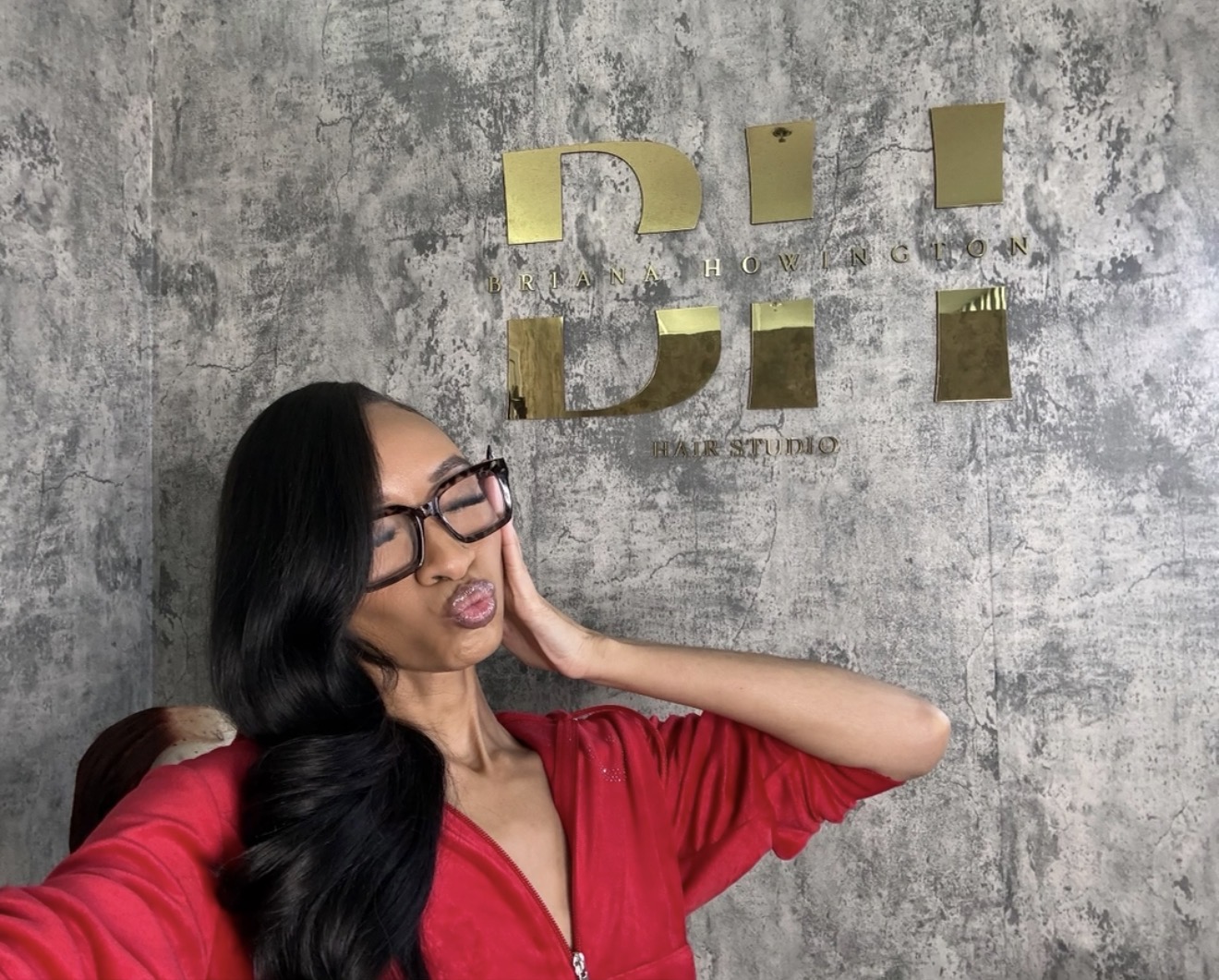 Woman with long black hair and glasses wearing a red top, posing with her hand on her face against a textured wall with gold letters.