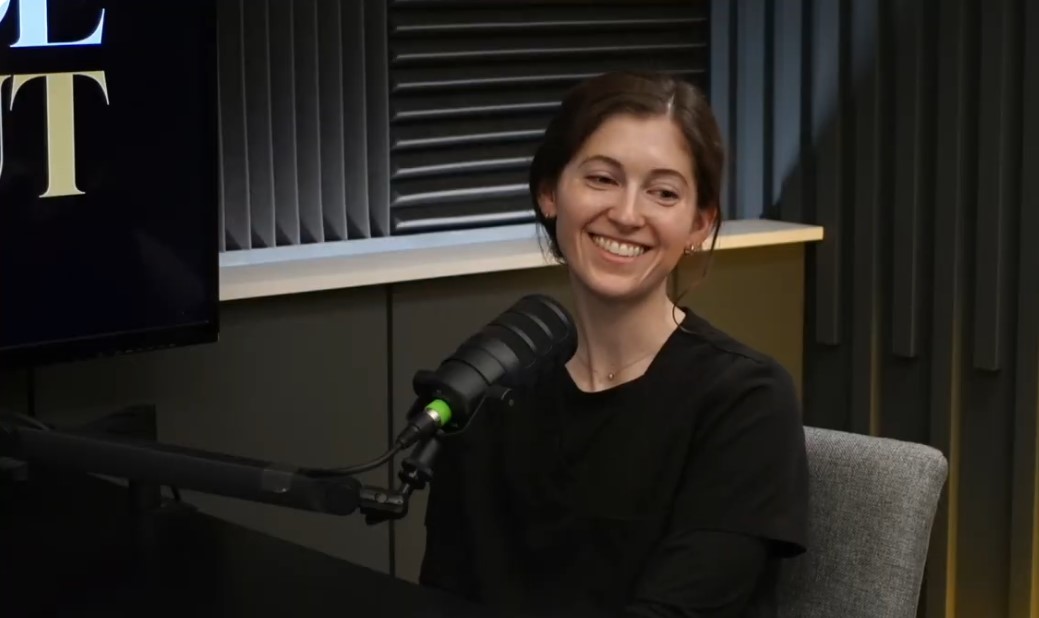 Young woman smiling, sitting at a desk with a microphone, in a recording studio, dark background.