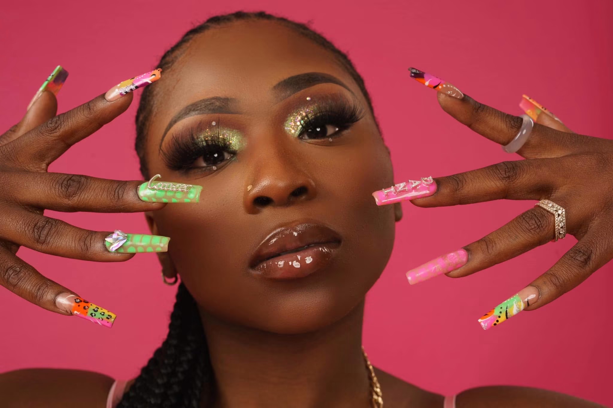 Close-up of a woman with long, decorated nails, makeup, and braided hair against a pink background.