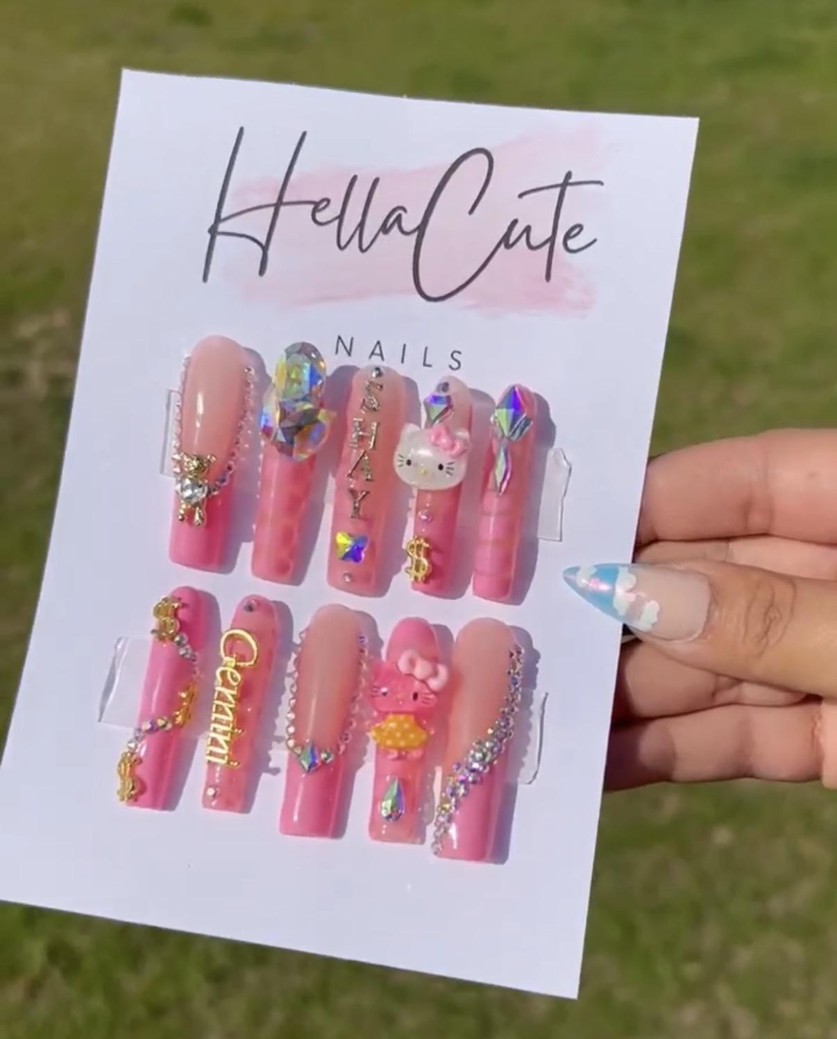 Hand holding a card with pink decorated nails and a unicorn nail art design, outdoors on grass.