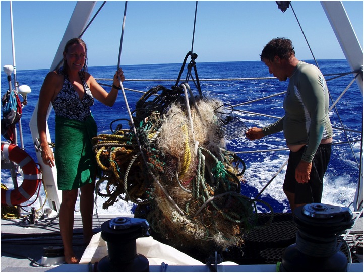 Two people on a boat with a large fish caught in fishing gear, ocean in background, clear sky.
