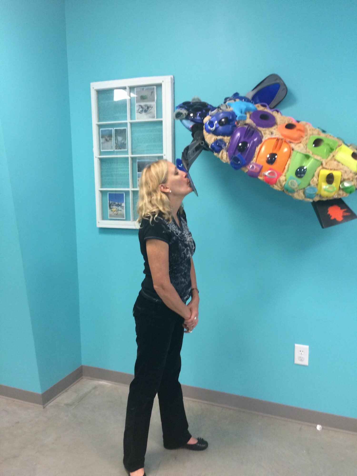 Woman with blonde hair standing sideways in front of a colorful fish-shaped balloon display against a teal wall.