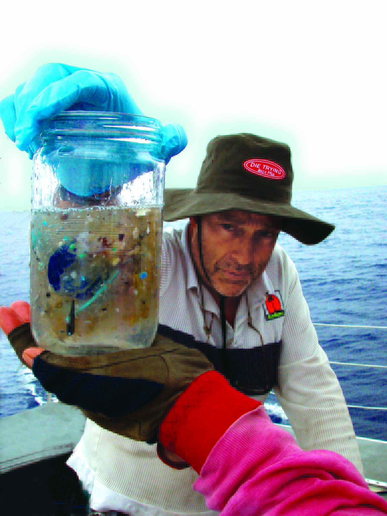 Person holding a jar with water and debris, wearing gloves and a hat, on a boat with water in the background.