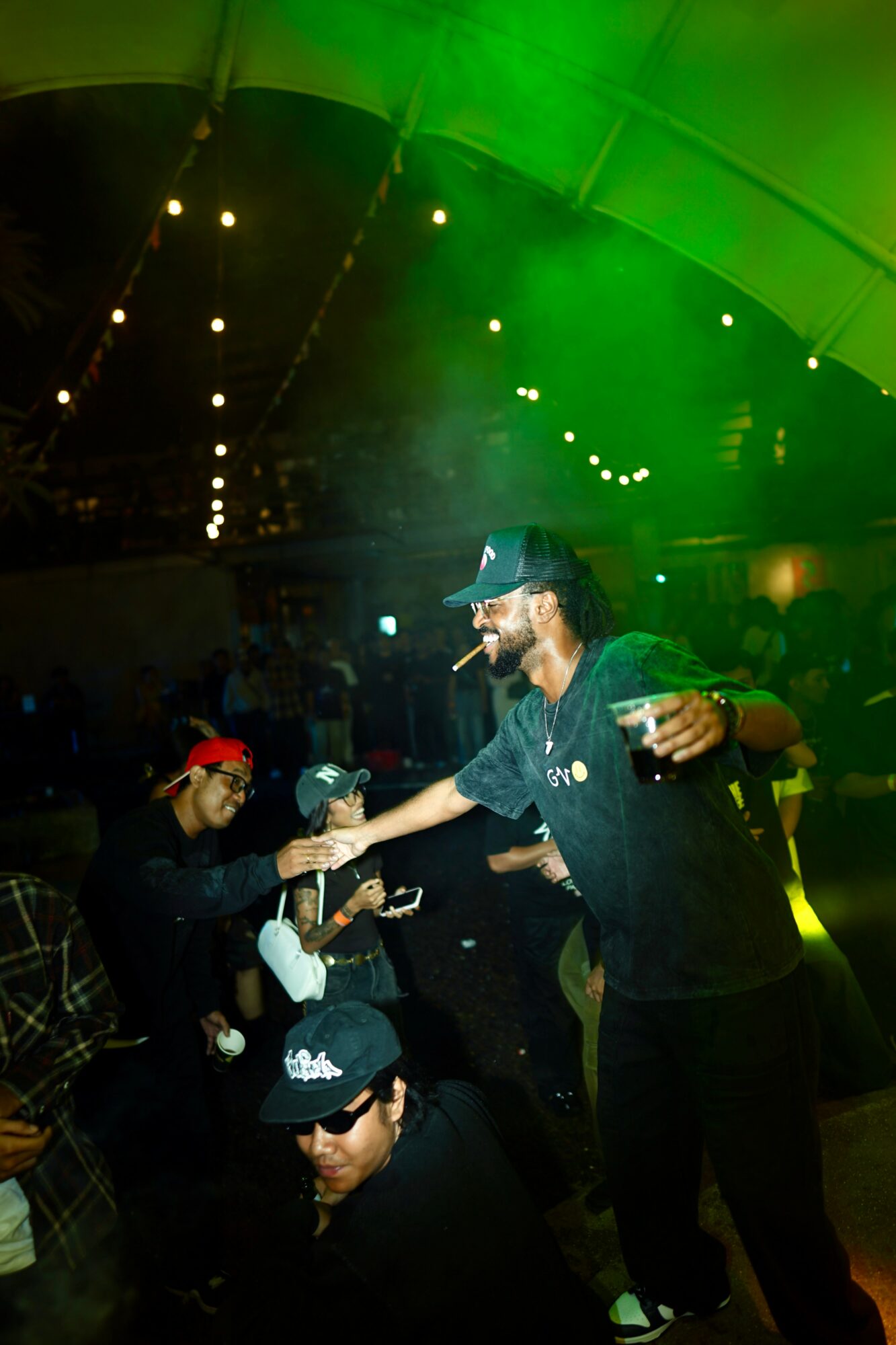 Person in black shirt and cap handing a drink to another person at a party with green lighting.