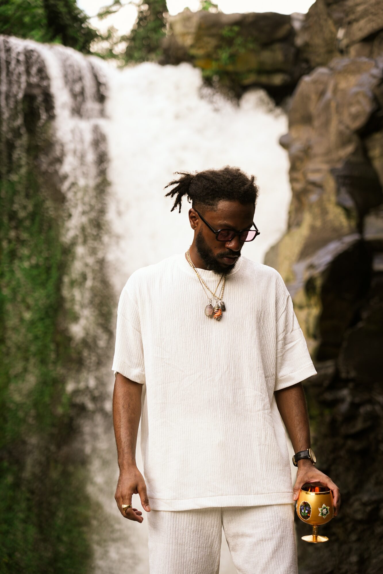 Man with dreadlocks and glasses stands in front of a waterfall, holding a small decorative object, wearing a white outfit.