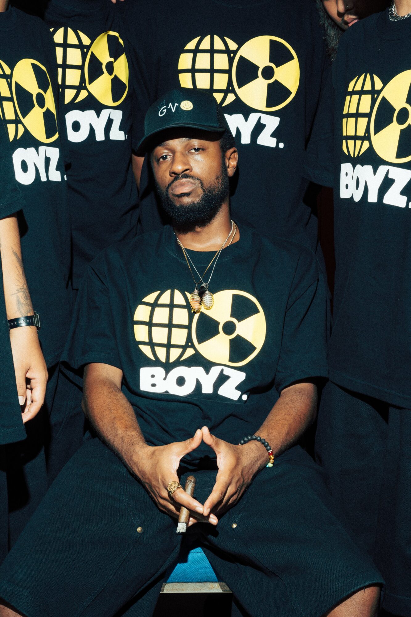 Man with beard and cap sitting with hands clasped, wearing a black T-shirt with a yellow and black logo, surrounded by others in similar shirts.