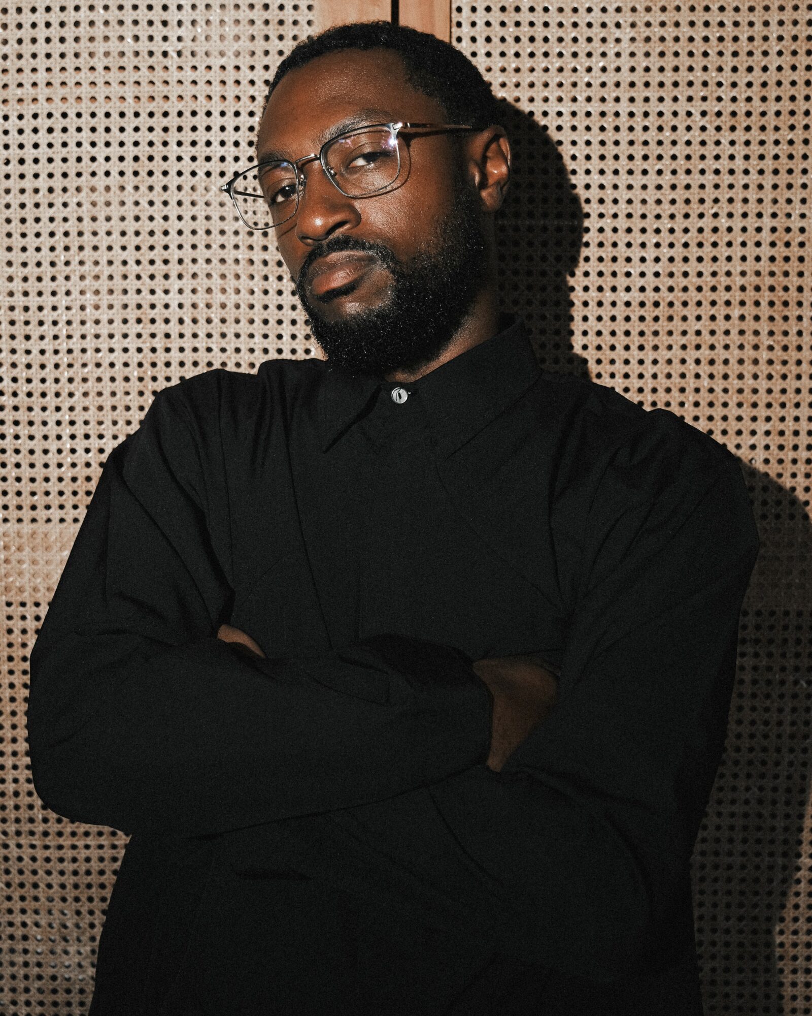 Man with glasses and beard wearing a black shirt, arms crossed, standing against a perforated wall.