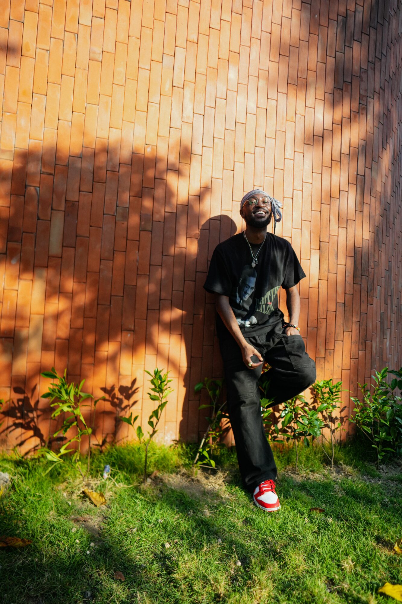Person leaning against a brick wall with shadows, standing on grass with plants, wearing black clothing and red shoes.