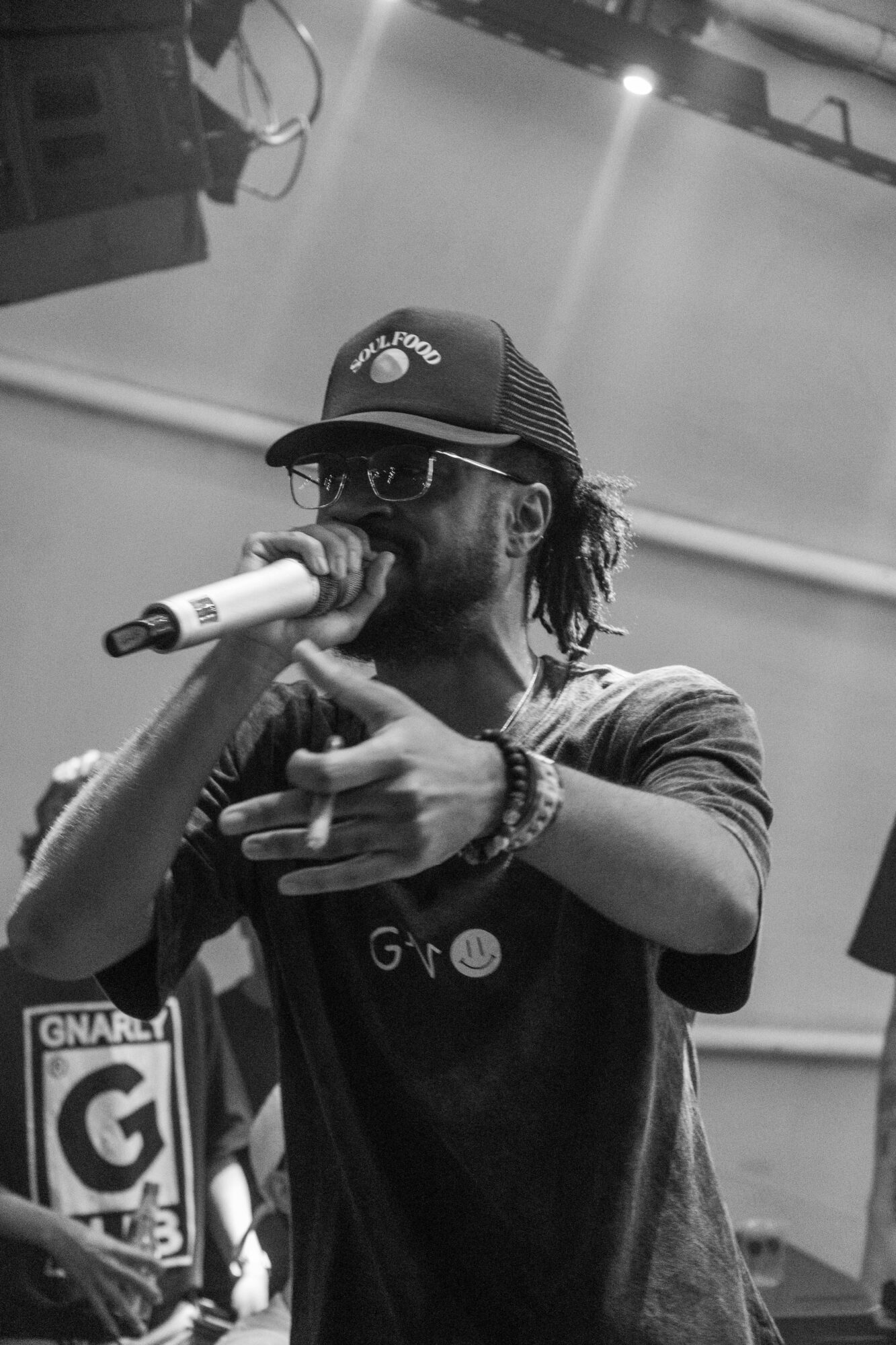 Person wearing a cap and glasses, holding a microphone, pointing forward, in a performance setting.