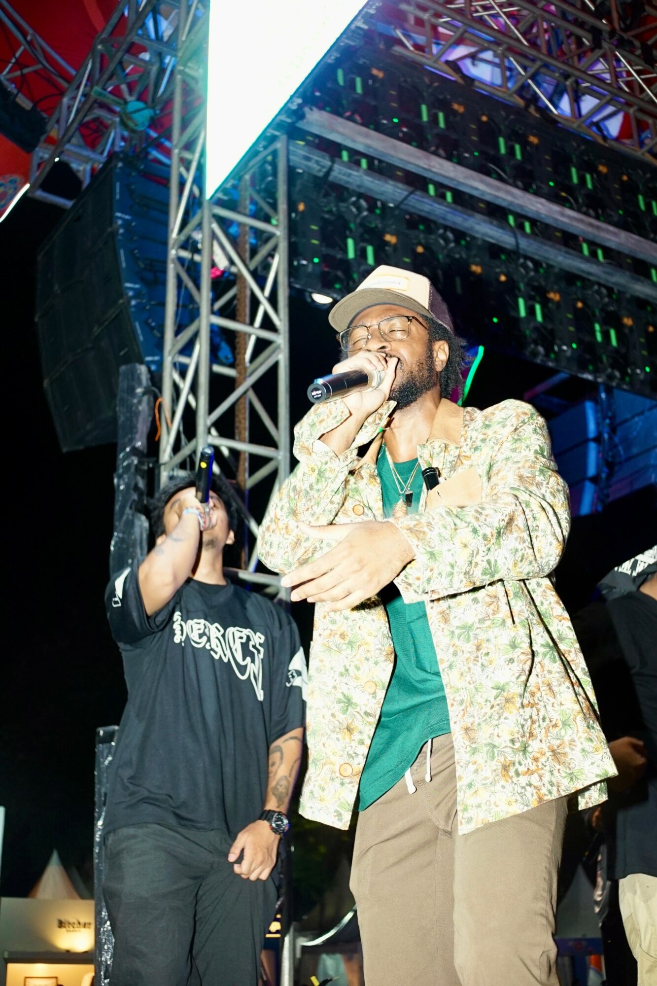 Person singing into a microphone on stage with colorful lights and metal scaffolding behind them.