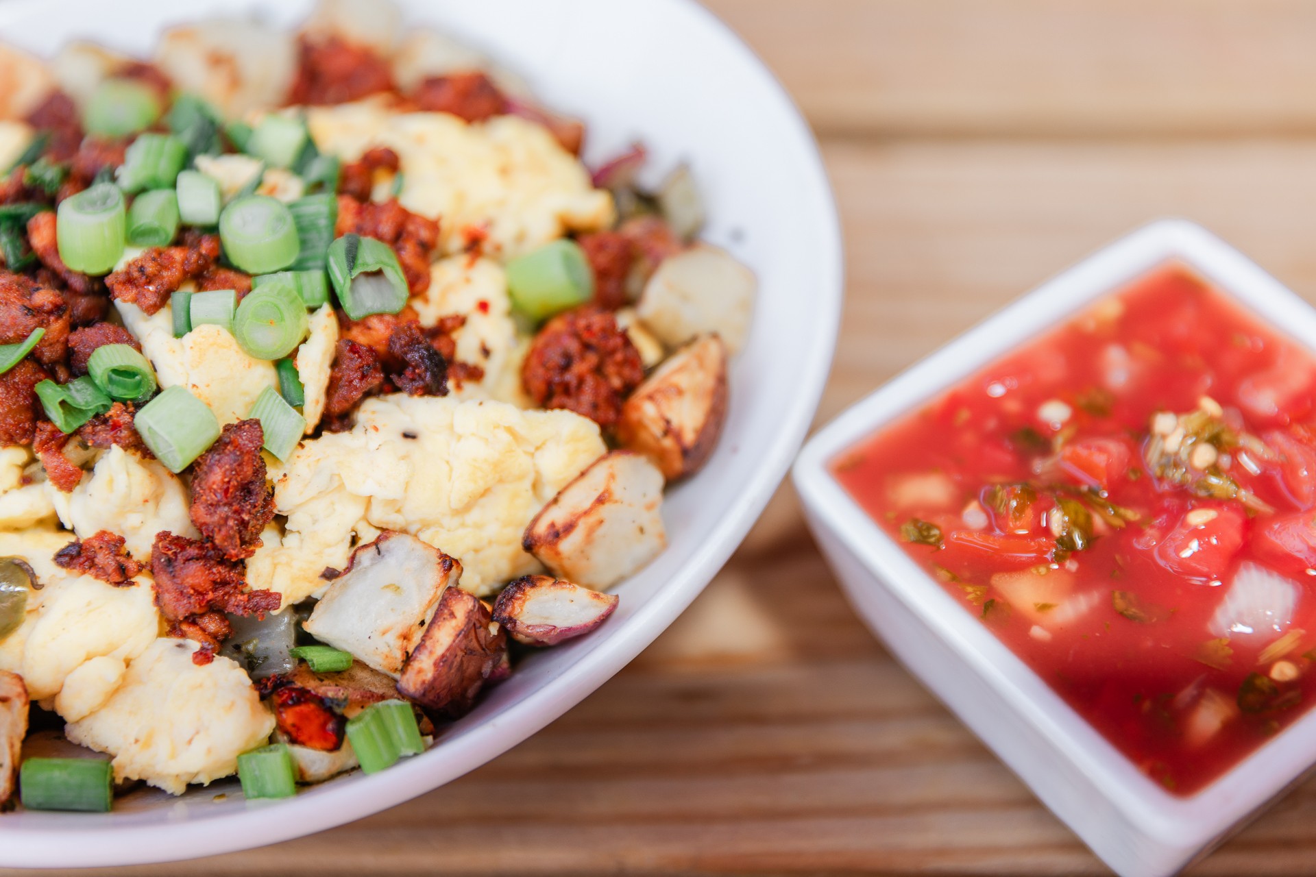 Bowl of fried chicken pieces with green onions and a side of tomato-based salsa in a small white dish on a wooden surface.
