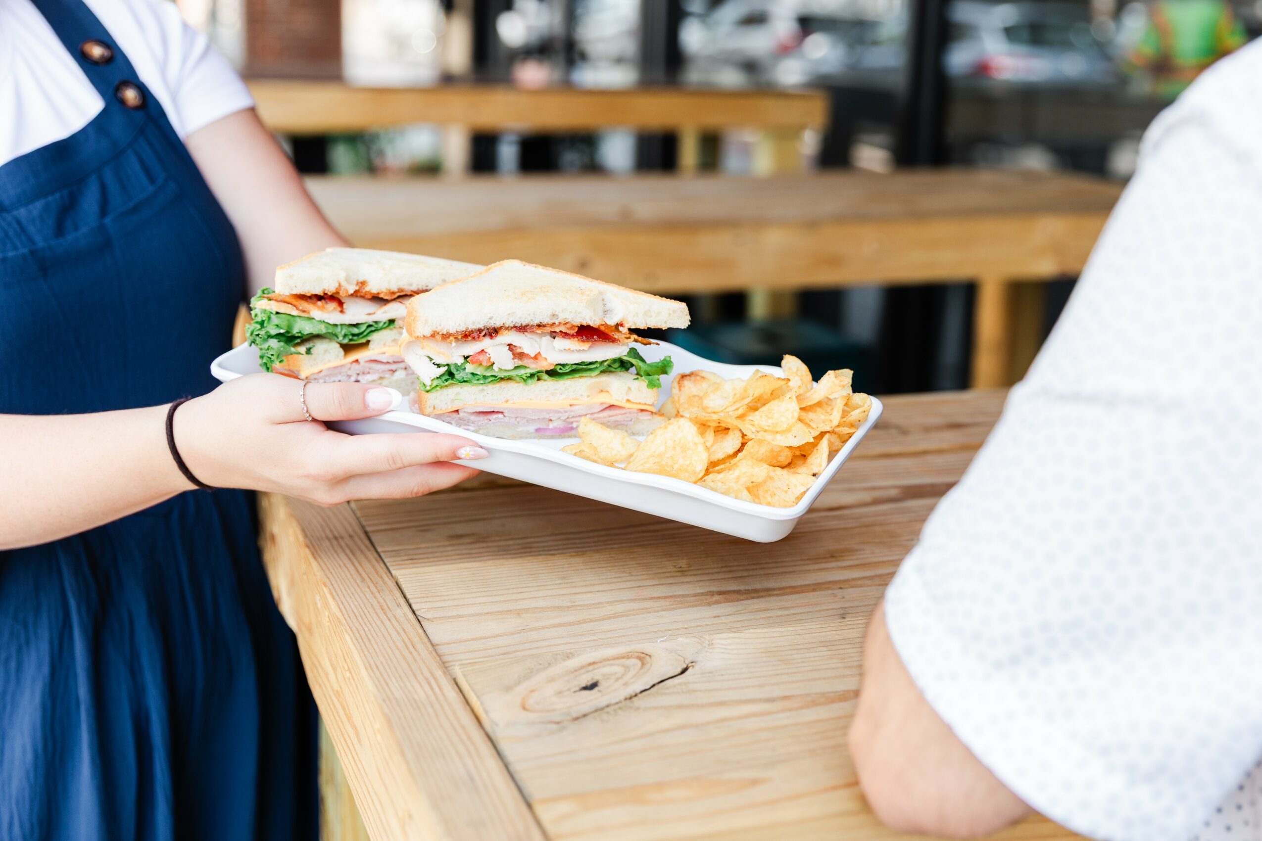Person holding a plate with a sandwich and chips, sitting at a wooden table in a casual setting.