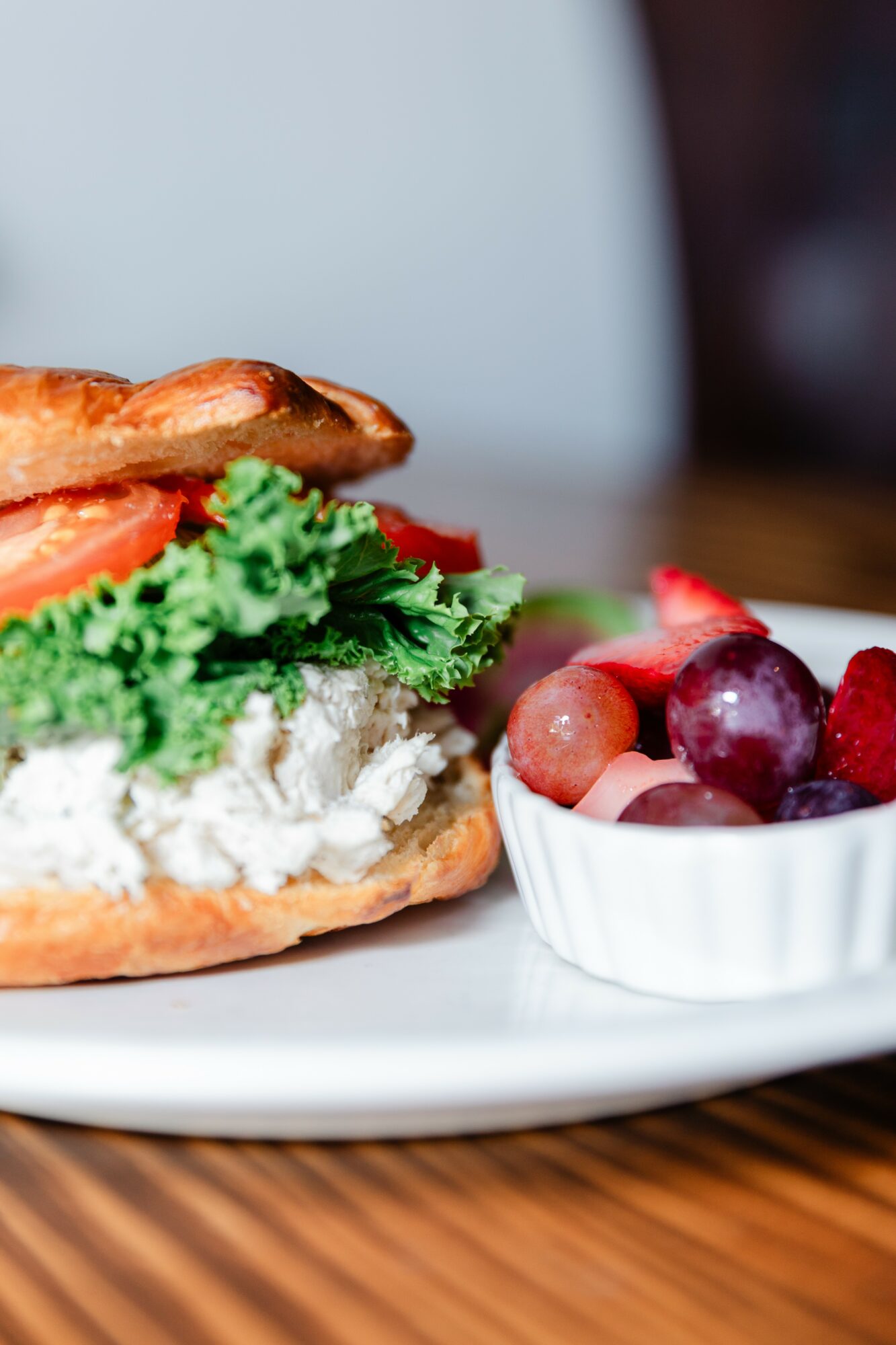 Sandwich with lettuce, tomato, and bacon on a bun, served with a bowl of mixed grapes on a white plate.