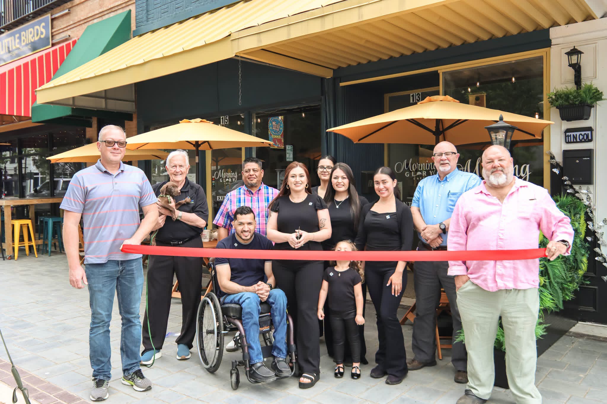 Group of nine people, including a person in wheelchair, standing outside a restaurant with yellow umbrellas, holding a red ribbon.