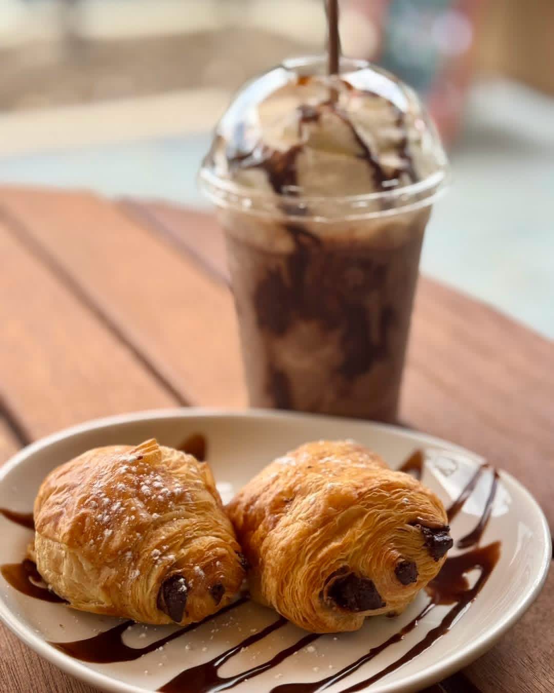 Two chocolate chip croissants on a white plate with chocolate drizzle, and a tall glass of iced chocolate drink in the background.