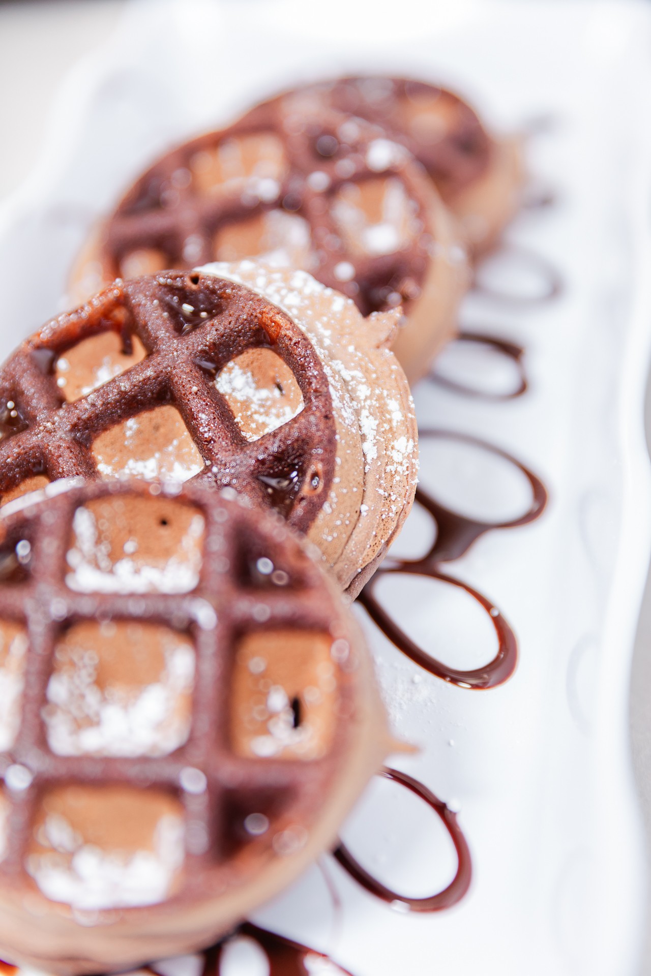 Three round waffles with chocolate drizzle and powdered sugar on a white plate, with some chocolate sauce on the side.