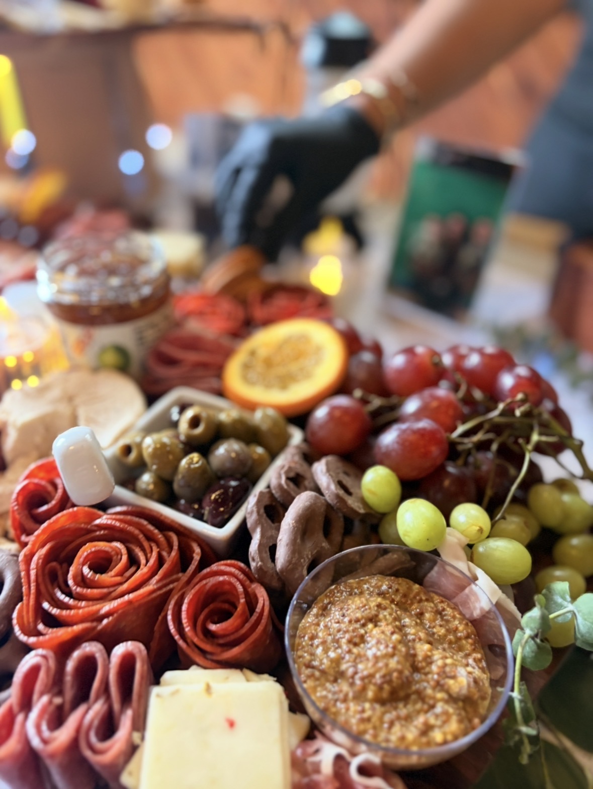 Assorted fruits, olives, and baked goods on a table with a person in the background wearing gloves.