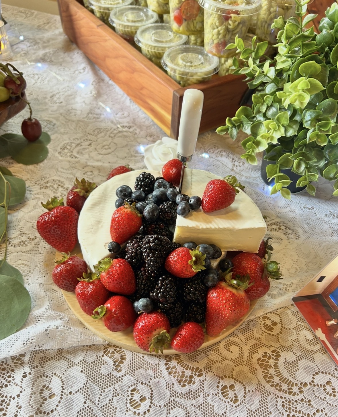 A cheese wheel topped with strawberries, blackberries, and blueberries, with a cheese knife inserted, on a lace tablecloth with plants and jars nearby.