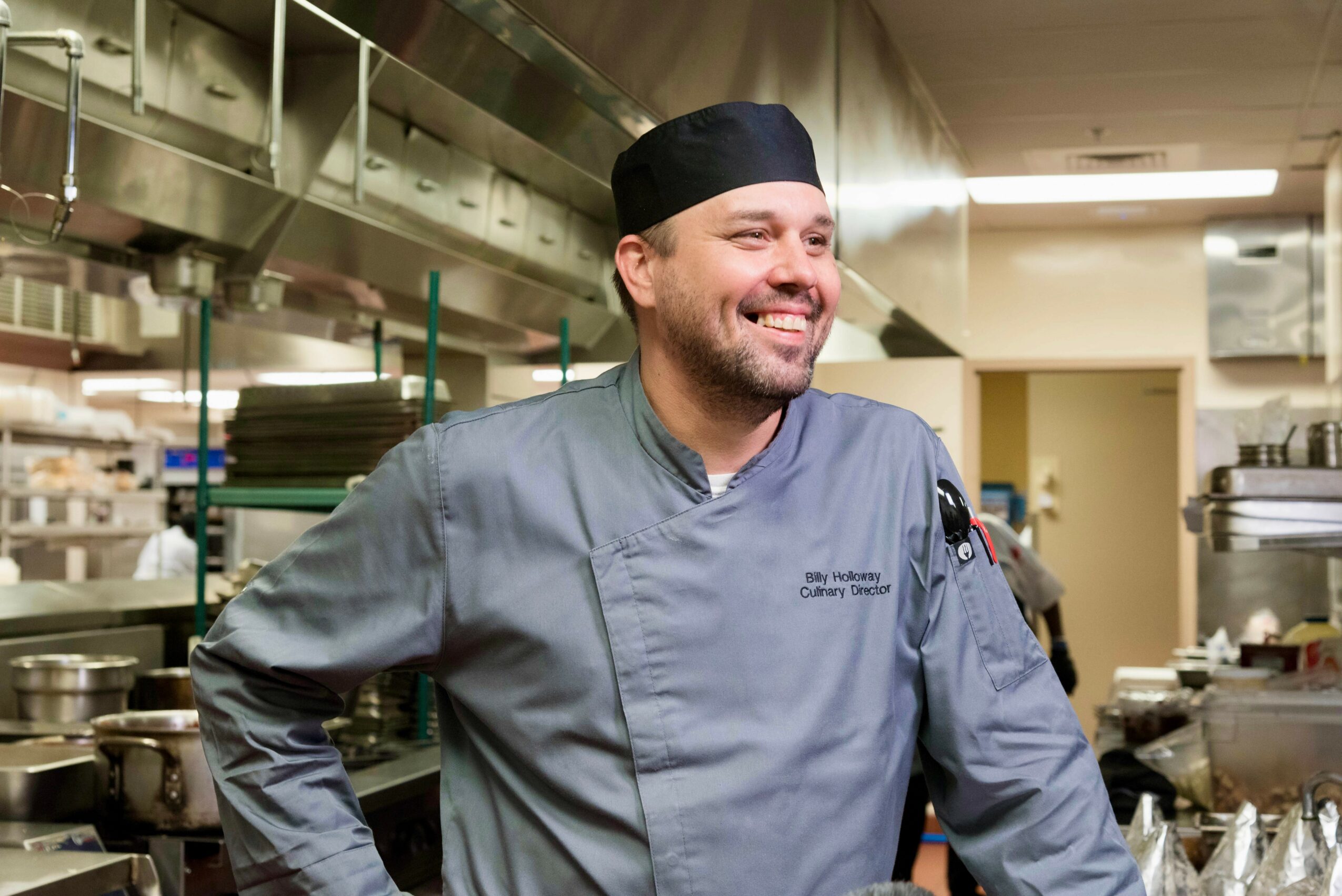 Smiling chef in gray uniform and black hat in a professional kitchen with stainless steel equipment.