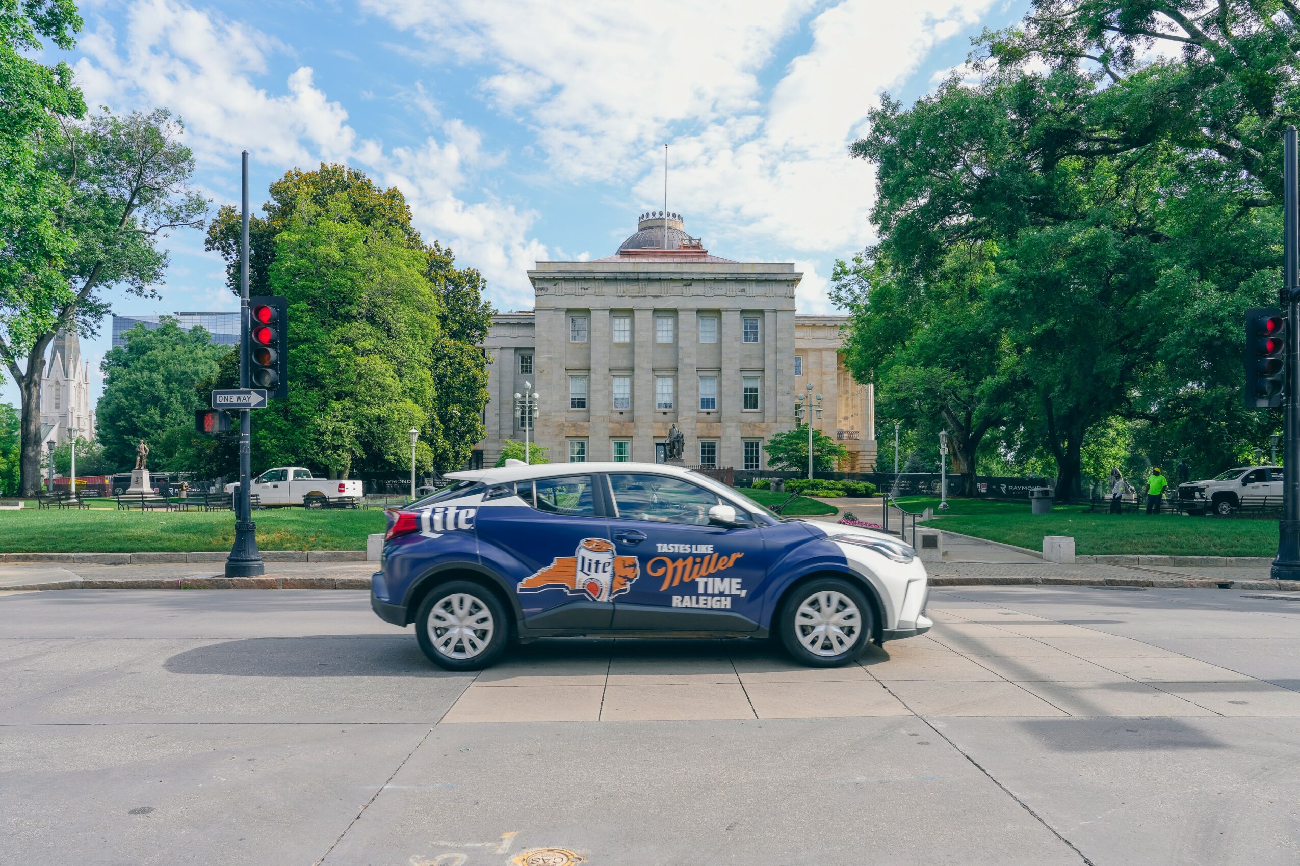 A white SUV with a logo and text on its side is parked on a city street in front of a large government building, trees, and a blue sky with clouds.