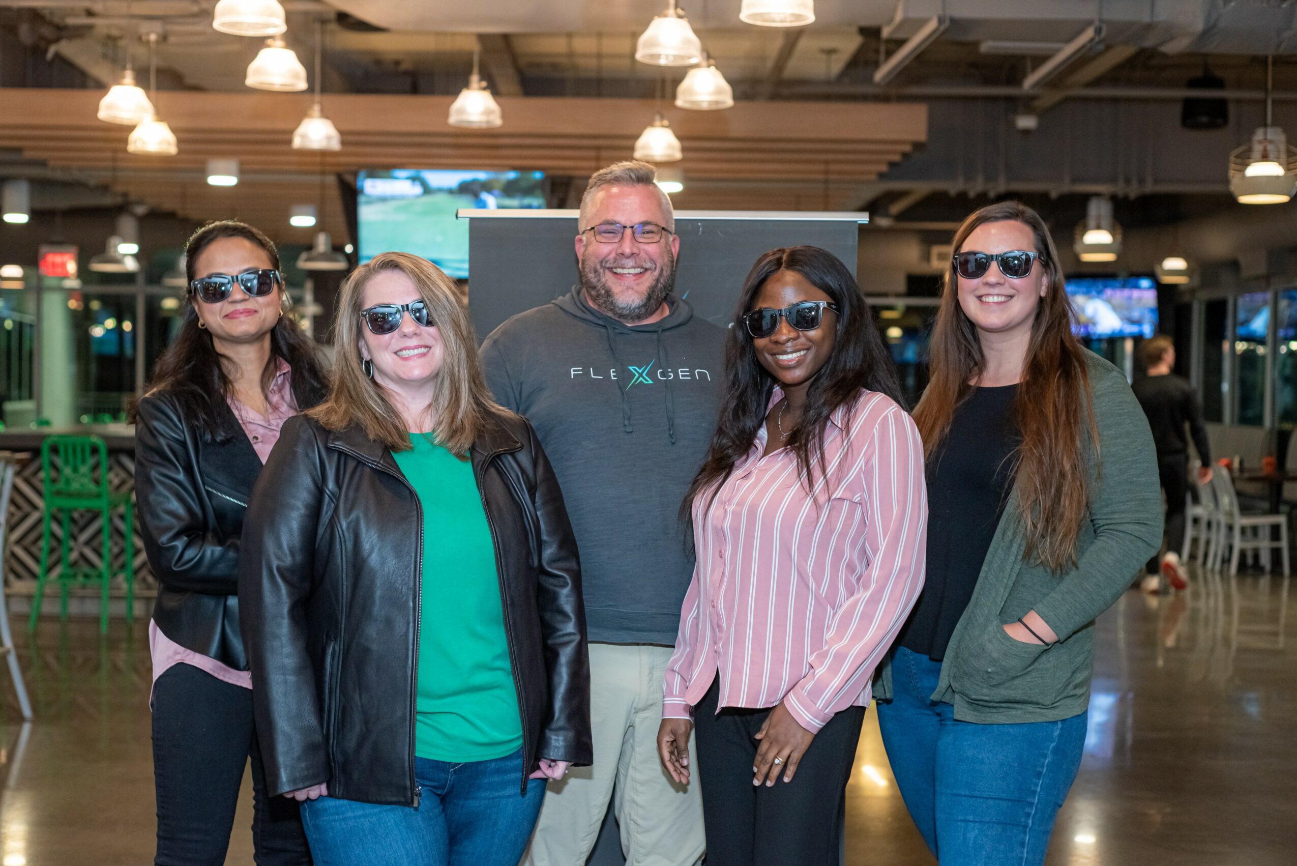Group of five people standing indoors, smiling, wearing casual clothing and sunglasses.