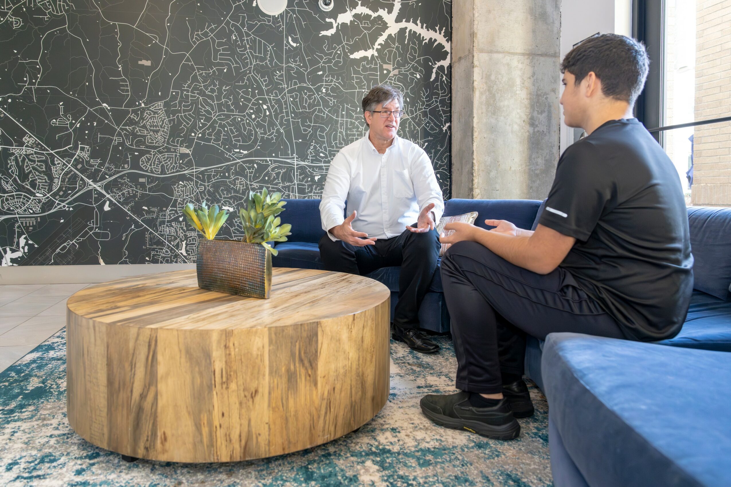 Two men sit on a blue sofa in a modern room, engaged in conversation, with a large map on the wall behind them.