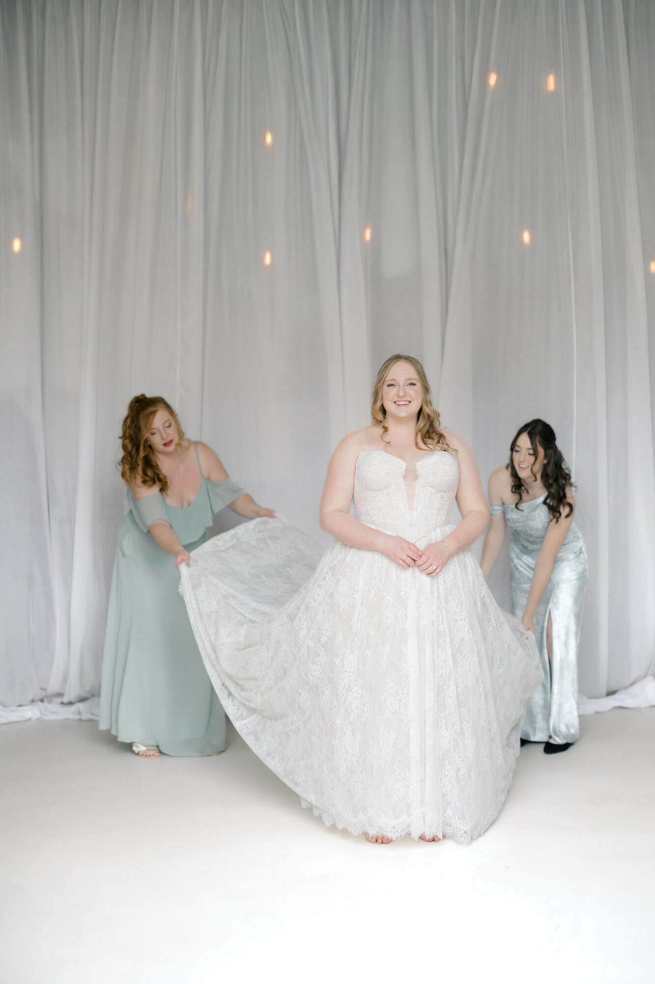 A bride in a white wedding dress stands between two bridesmaids in light blue dresses, in front of a white curtain backdrop with small lights.