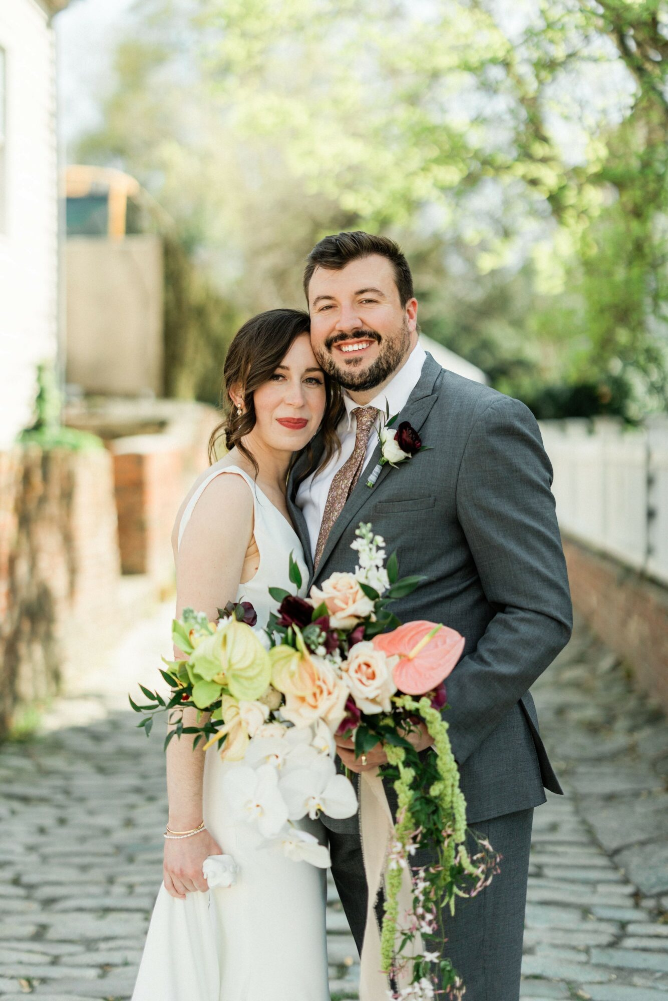 A couple in wedding attire outdoors, smiling, with trees and a pathway in the background.