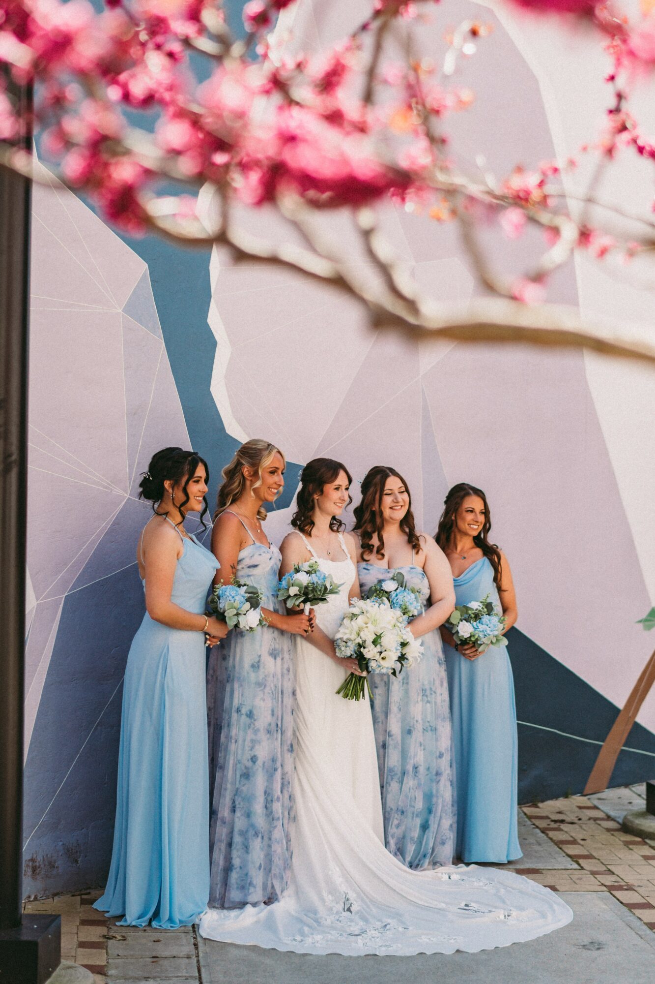 Five women in wedding dresses and blue bridesmaid dresses holding bouquets, standing outdoors against a geometric wall, with pink blossoms in the foreground.