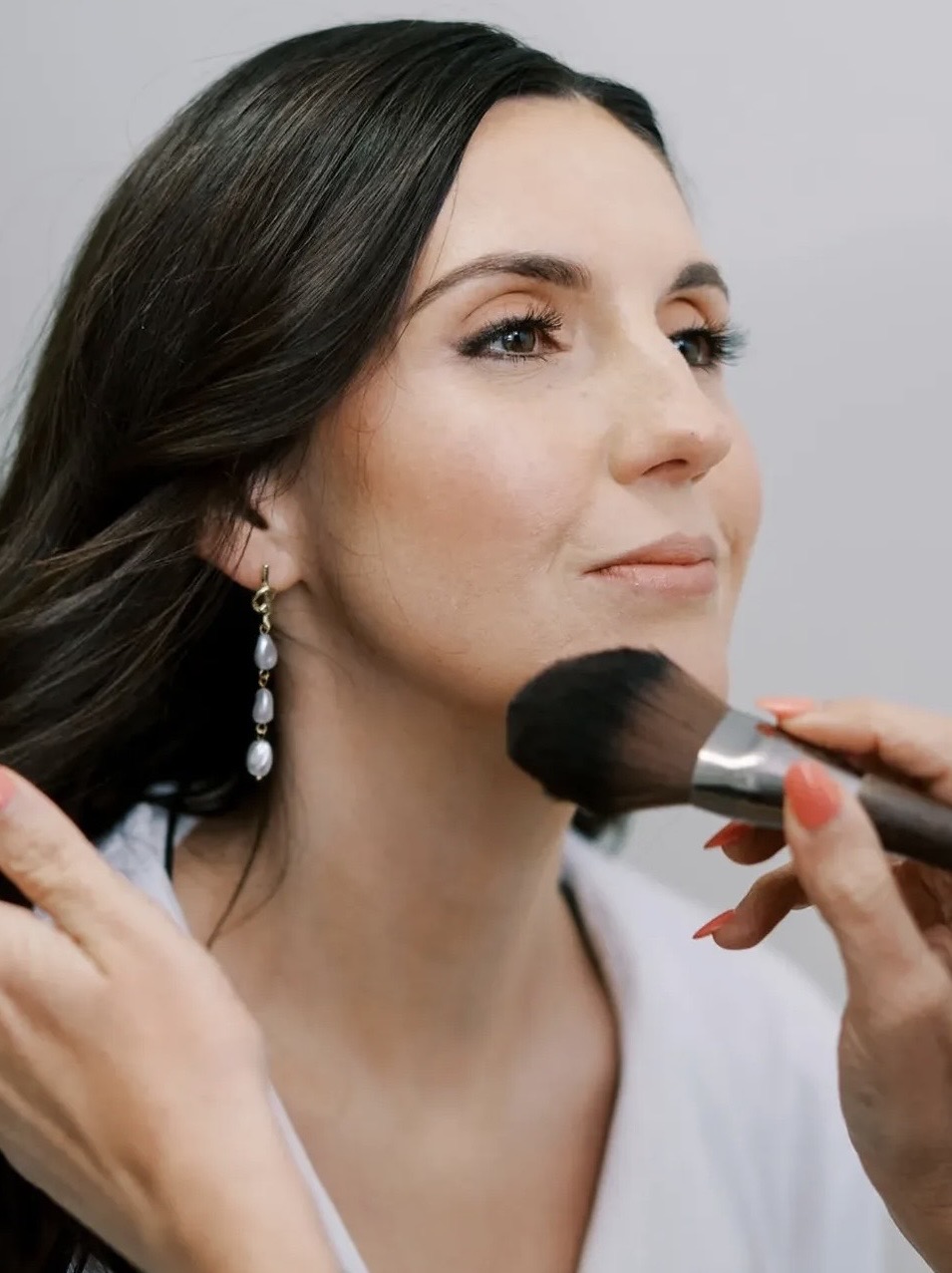 Woman with dark hair and earrings having makeup applied with a brush, facing slightly to the right.