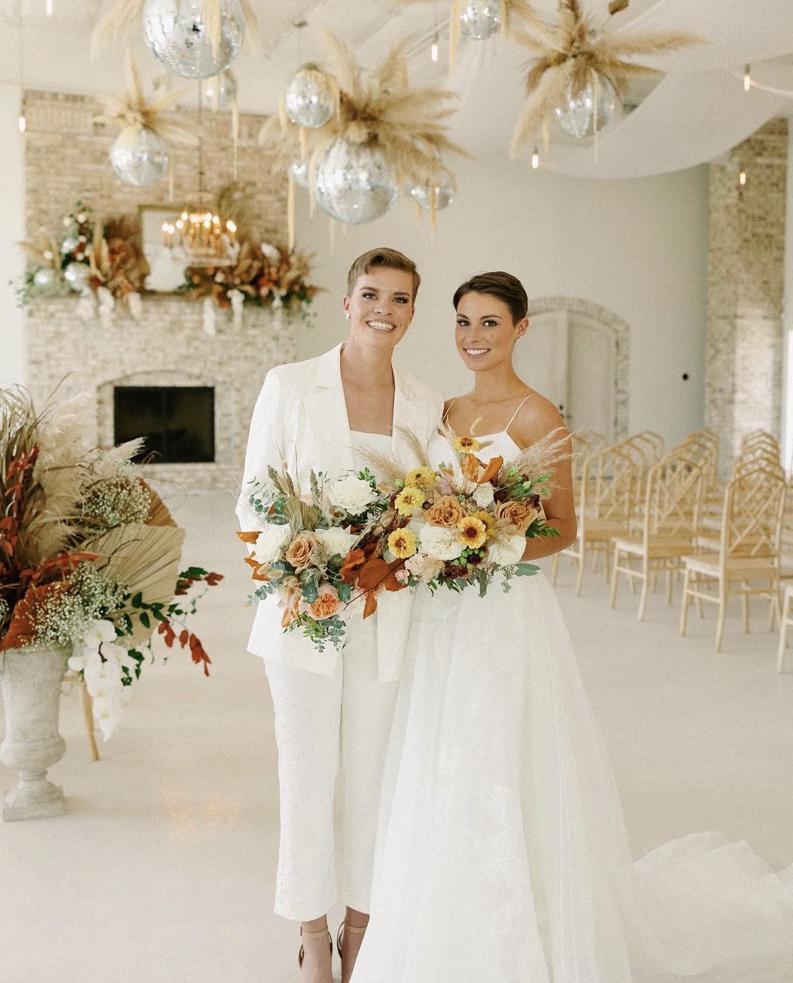 Two women in wedding dresses holding bouquets, smiling indoors with decorated background and chairs.