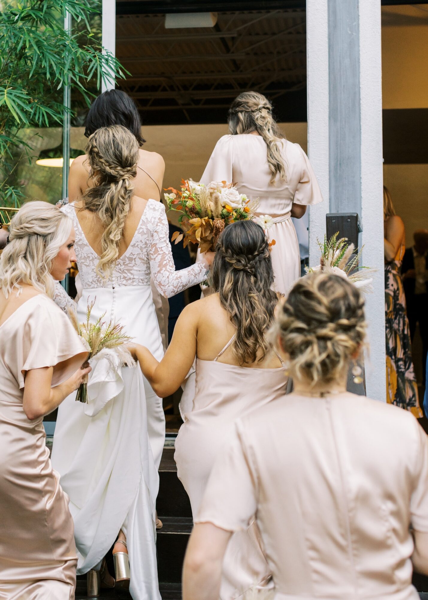 Group of women in dresses standing near a doorway, some holding flowers, with greenery and a building interior visible.