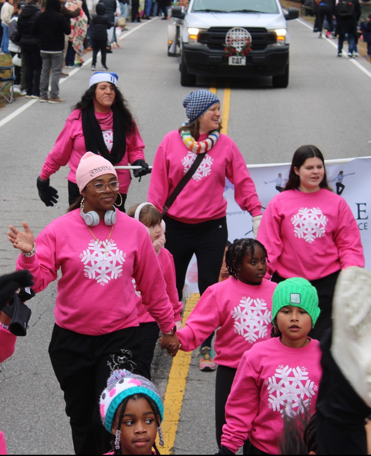 Group of people walking on a street during a parade, wearing pink sweatshirts with white snowflake designs, some holding hands.