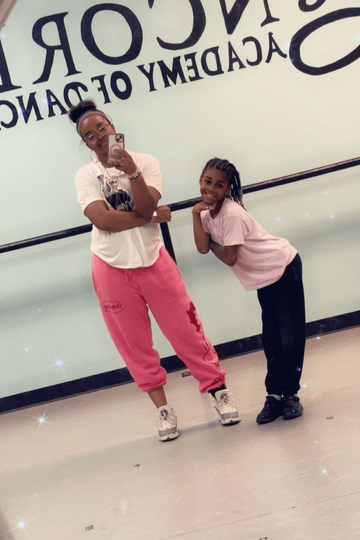 Two young girls pose playfully in front of a wall with text, one with hand on chin, smiling, and the other leaning on her shoulder.
