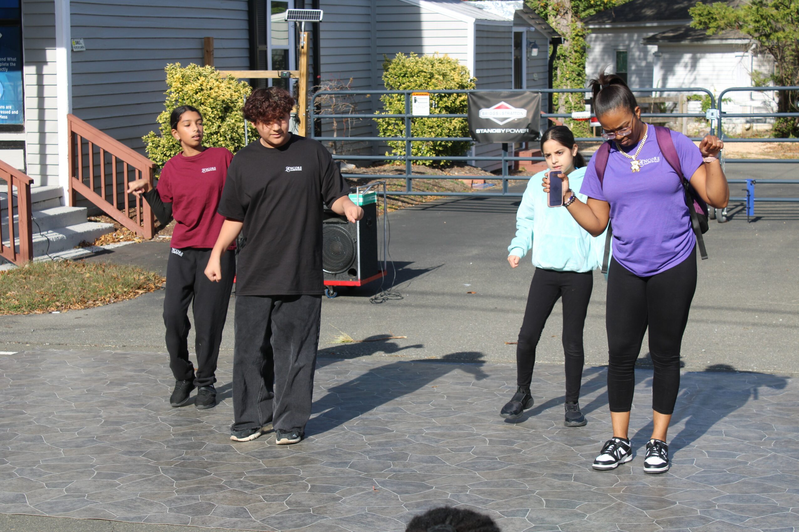 Four people standing outdoors on a paved area, two women and two men, engaged in conversation, with buildings and trees in background.